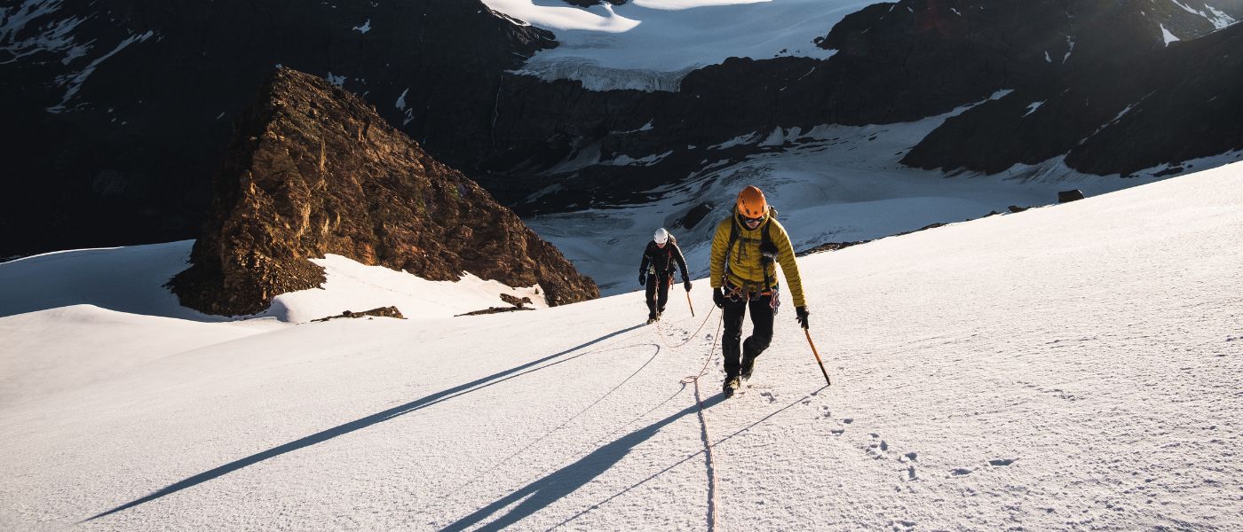 Eine Gruppe von Menschen erklimmt einen verschneiten Berg, einige tragen gelbe Jacken und Helme, ausgestattet mit Wanderstöcken.