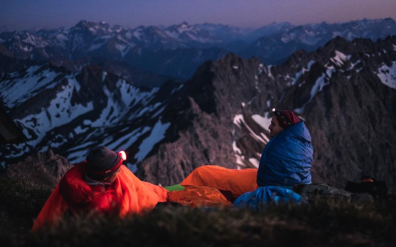 Zwei Personen sitzen auf einem Berg mit Schlafsäcken, eine trägt eine rote Jacke. Szene vermittelt Outdoor-Abenteuer, passend zu alpinloacker.com Produkten.