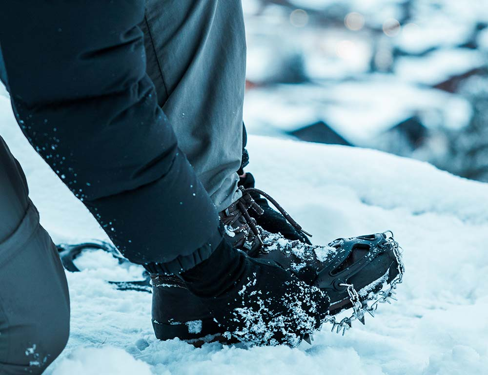 Person trägt schwarze Schuhe mit Spikes im Schnee, ideal für winterliche Outdoor-Aktivitäten. Perfekt für Alpinloacker-Kunden, die robuste Winterausrüstung suchen.