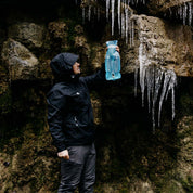 Person in dunkler Herren 3 Lagen Hardshell Regenjacke neben Felswand, inspiziert Wasser mit blauem Reservoir. Ideal für Outdoor-Aktivitäten dank wasser- und winddichter Eigenschaften.