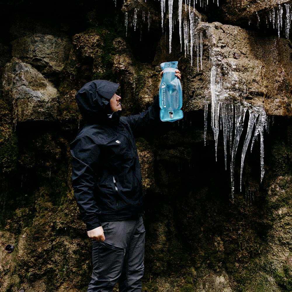Person in dunkler Herren 3 Lagen Hardshell Regenjacke neben Felswand, inspiziert Wasser mit blauem Reservoir. Ideal für Outdoor-Aktivitäten dank wasser- und winddichter Eigenschaften.