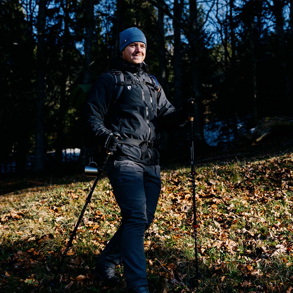 Person beim Wandern im Wald, trägt Herren 3 Lagen Hardshell Regenjacke von Alpin Loacker, ausgestattet mit Trekkingstöcken und Rucksack, ideal für Outdoor-Aktivitäten.