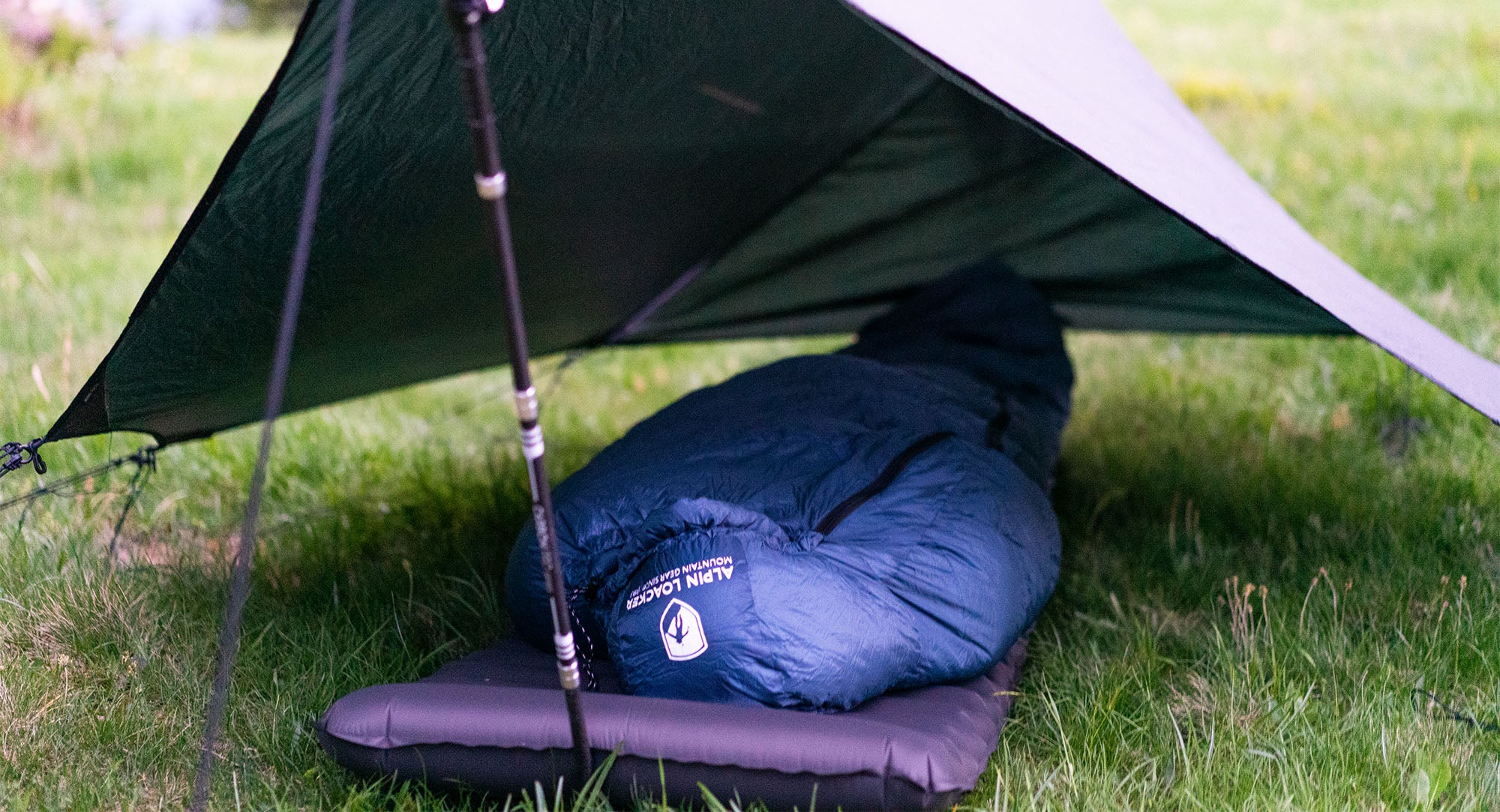 Ein blauer Schlafsack mit Logo liegt unter einem Zelt auf Gras, ideal für Outdoor-Campingausflüge.
