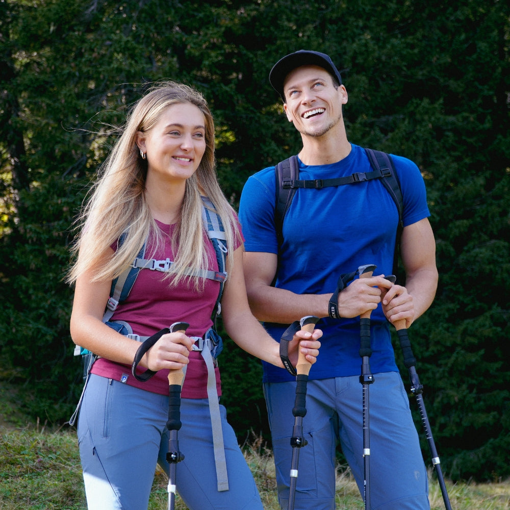 Ein Mann und eine Frau mit Wanderstöcken, beide tragen Outdoor-Kleidung und Rucksäcke, im Freien lächelnd, passend für Outdoor-Aktivitäten von alpinloacker.com.