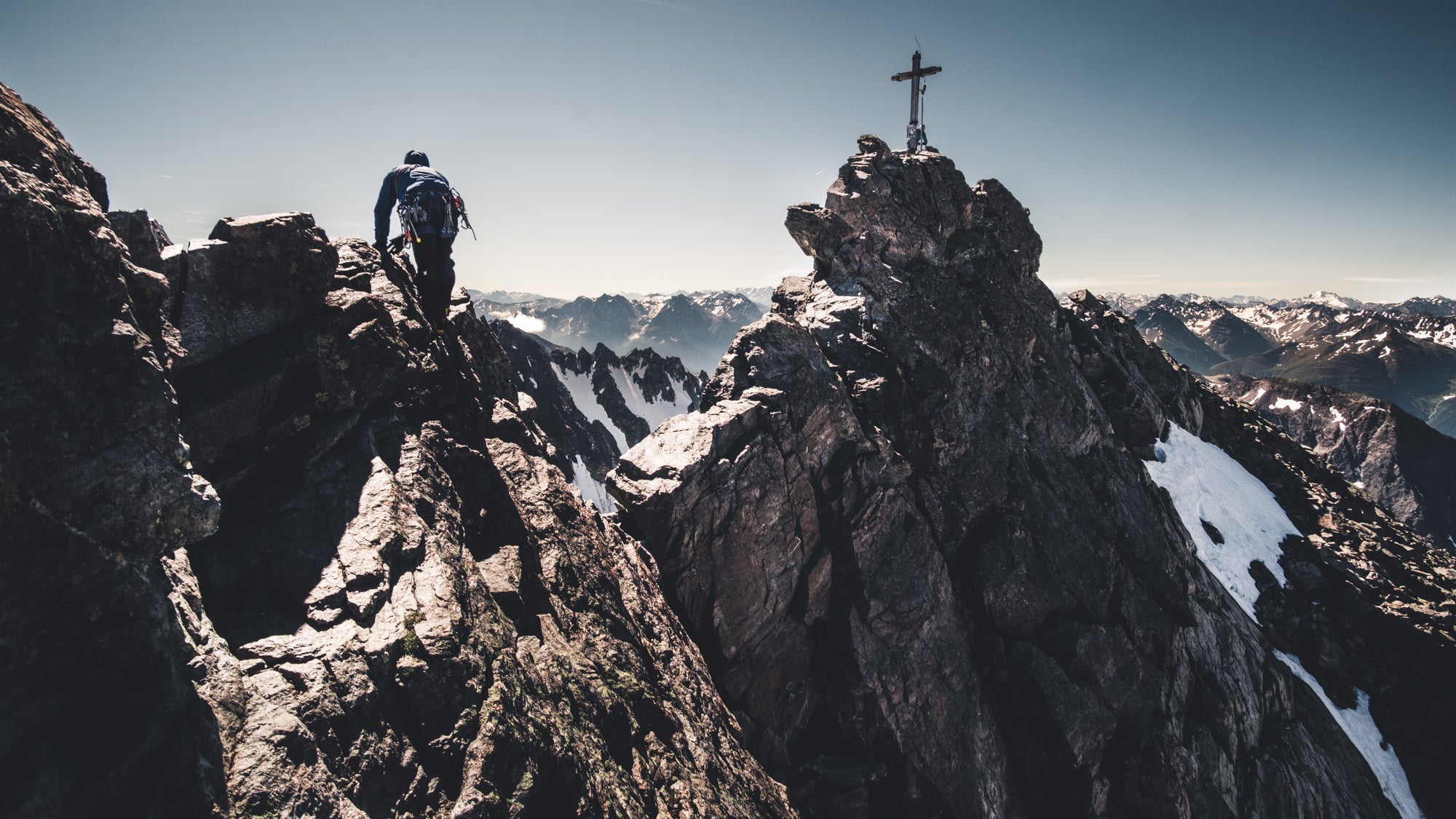 Eine Person mit Rucksack steht auf einem verschneiten Berggipfel neben einem Kreuz, umgeben von felsiger Landschaft.