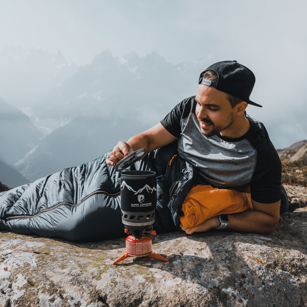 Ein Mann liegt auf einem Felsen neben einem Gaskocher, trägt Outdoor-Kleidung und blickt in die Natur. Ideal für Alpinloacker-Abenteuer.