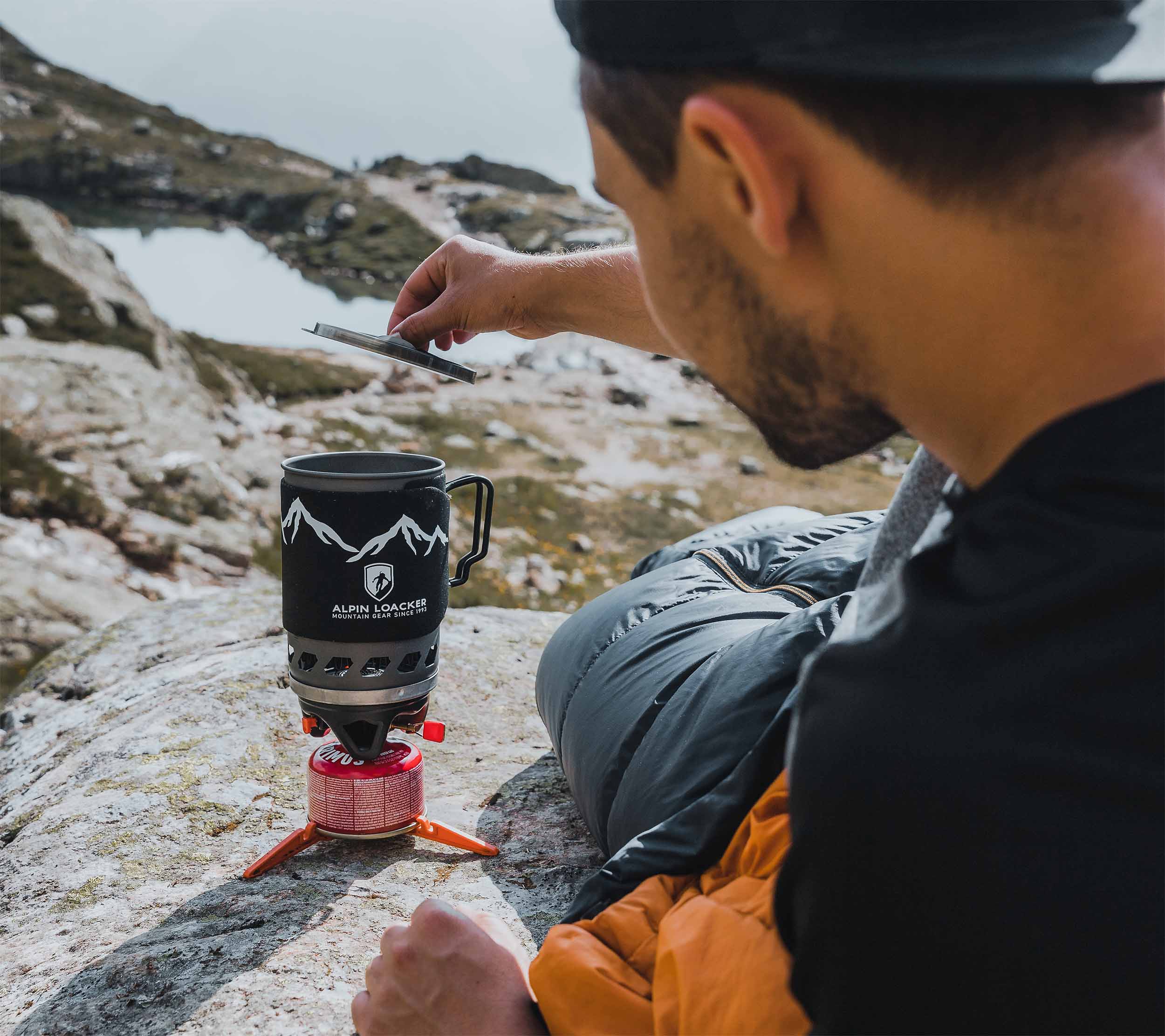 Ein Mann sitzt auf einem Felsen und beobachtet einen Topf auf einem Gaskocher, möglicherweise während einer Outdoor-Expedition.