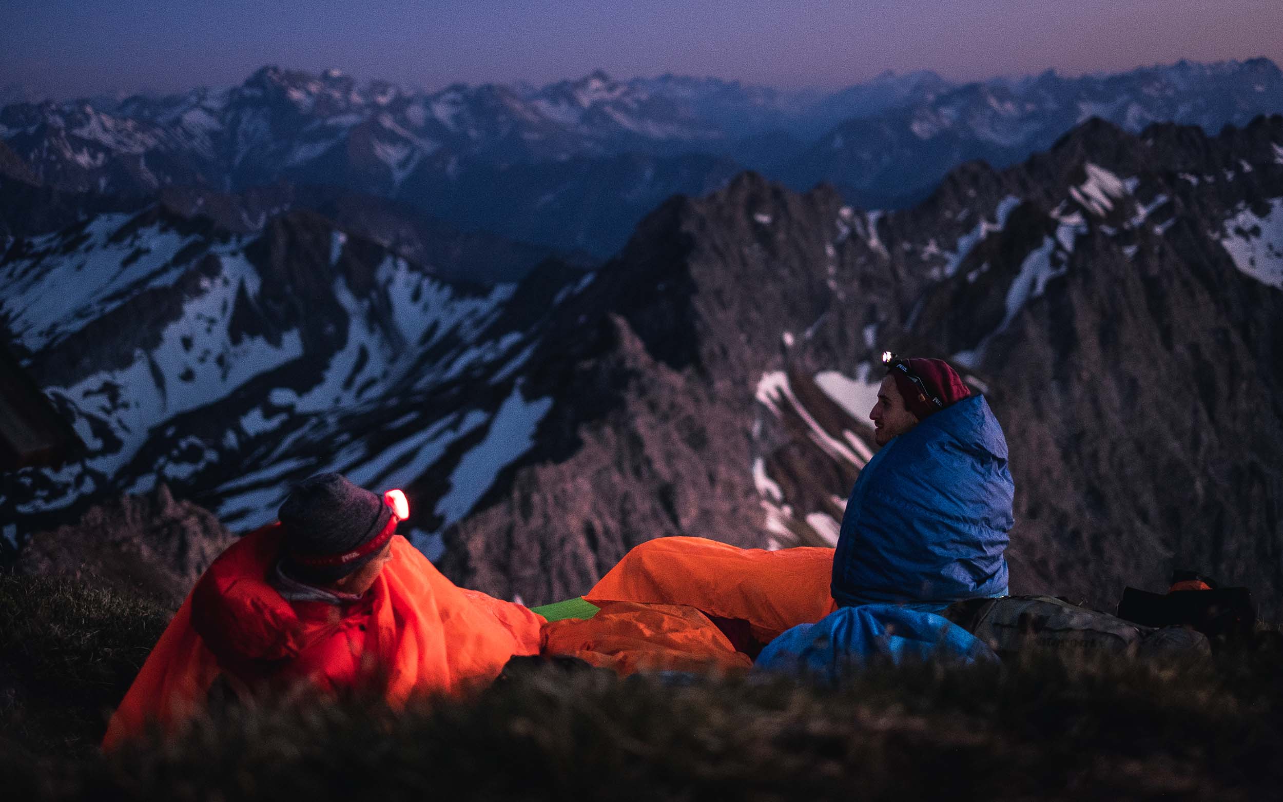 Zwei Personen in Schlafsäcken auf einem Berggipfel, mit Blick auf die Berglandschaft. Ideal für Outdoor-Aktivitäten und Produkte von alpinloacker.com.