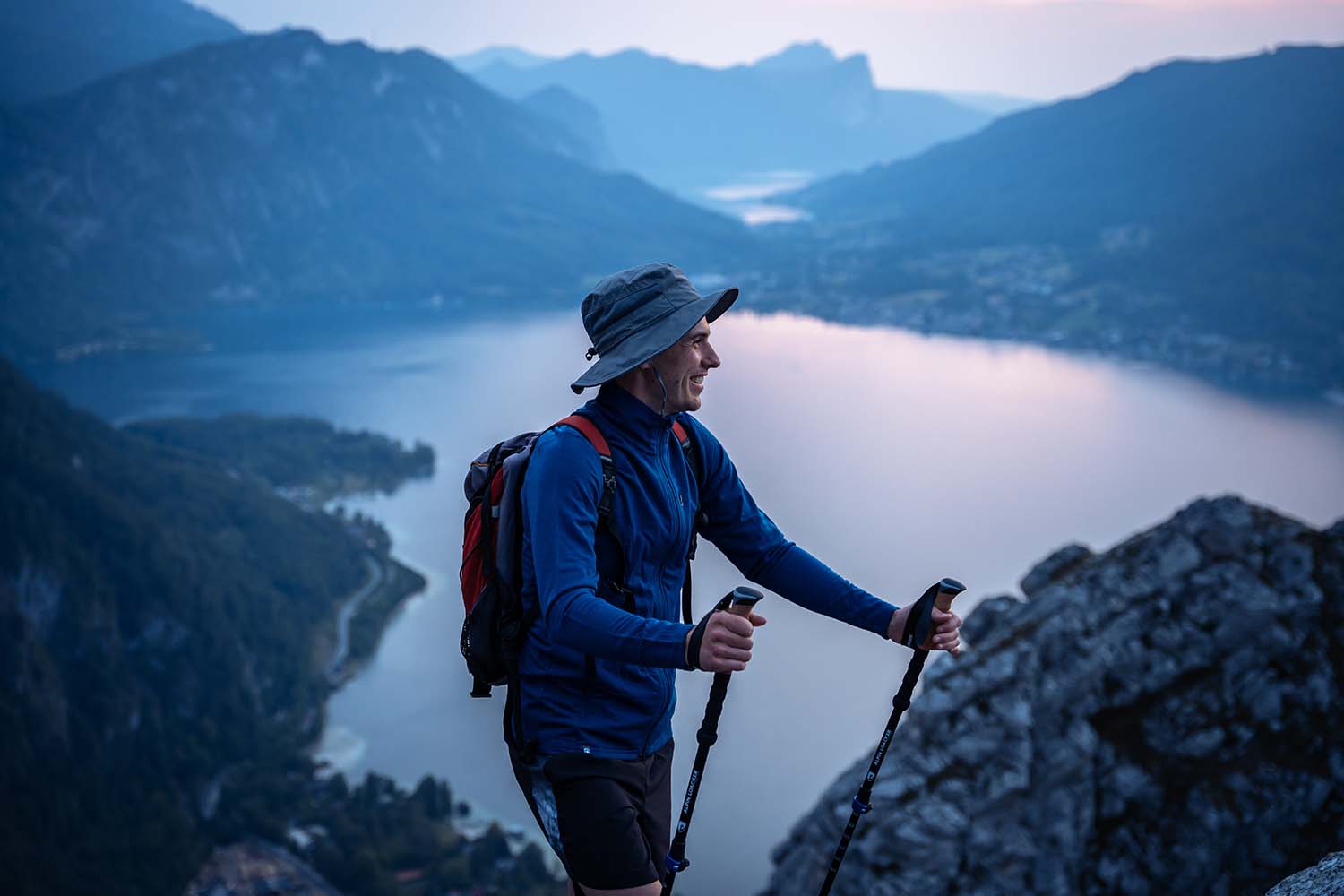 Ein Mann mit Hut und Rucksack steht auf einem Berg und hält Wanderstöcke, ideal für Outdoor-Abenteuer und Ausrüstung von alpinloacker.com.
