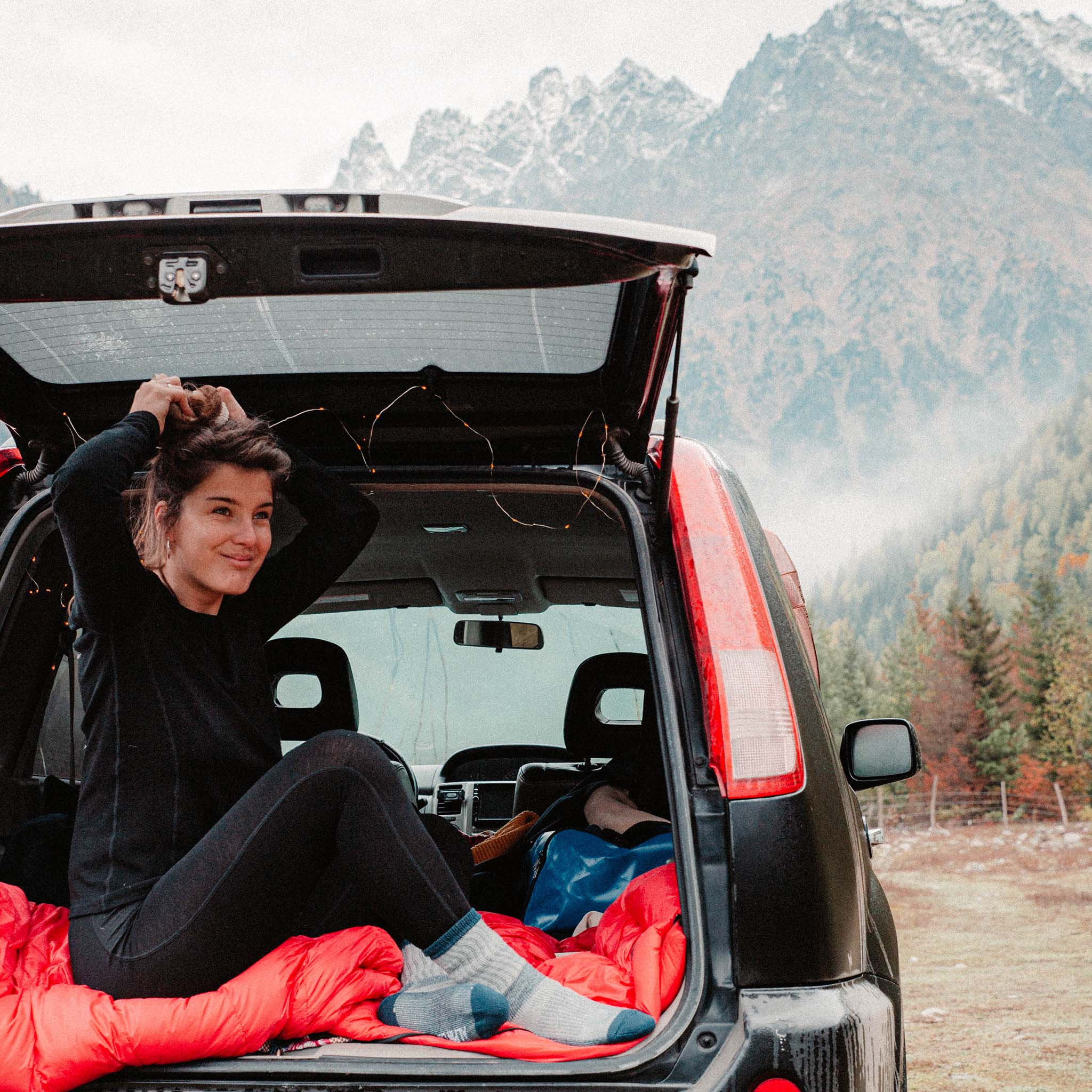 Eine Frau sitzt im Fond eines Autos, umgeben von Outdoor-Ausrüstung, passend für Aktivitäten wie Wandern und Camping.