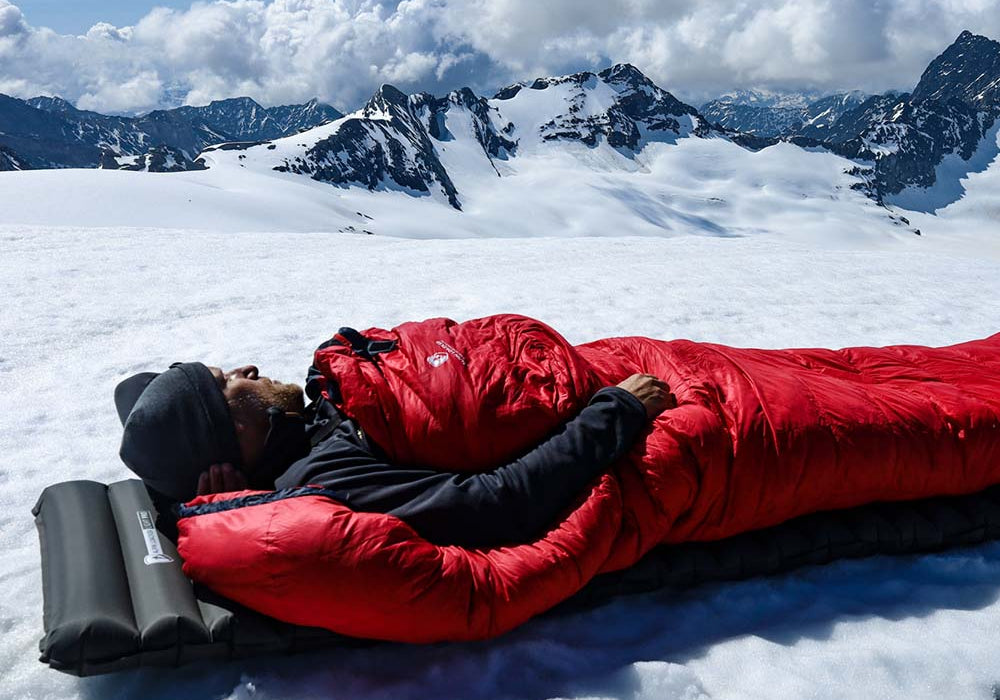 Ein Mann schläft in einem Schlafsack auf Schnee, vor einer verschneiten Berglandschaft unter wolkigem Himmel.