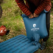 Person hält blaue, leichte Isomatte für 4 Jahreszeiten von Alpin Loacker, ideal für Outdoor-Abenteuer und effektive Isolation. Liegt auf Gras.