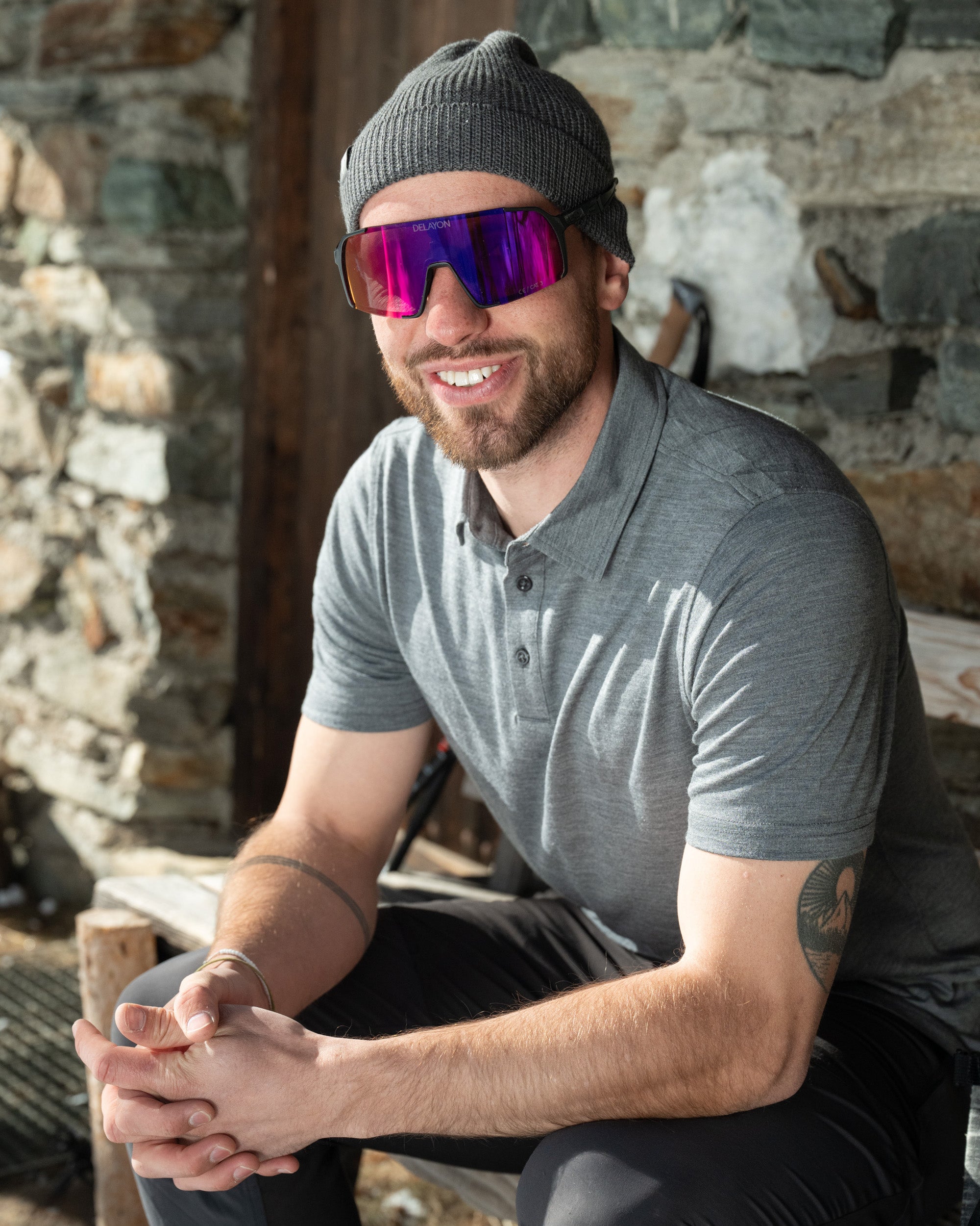 Model sitzt vor Hütte in grauem Merino Coolmax Polo Shirt - perfekt für Outdoor Aktivitäten