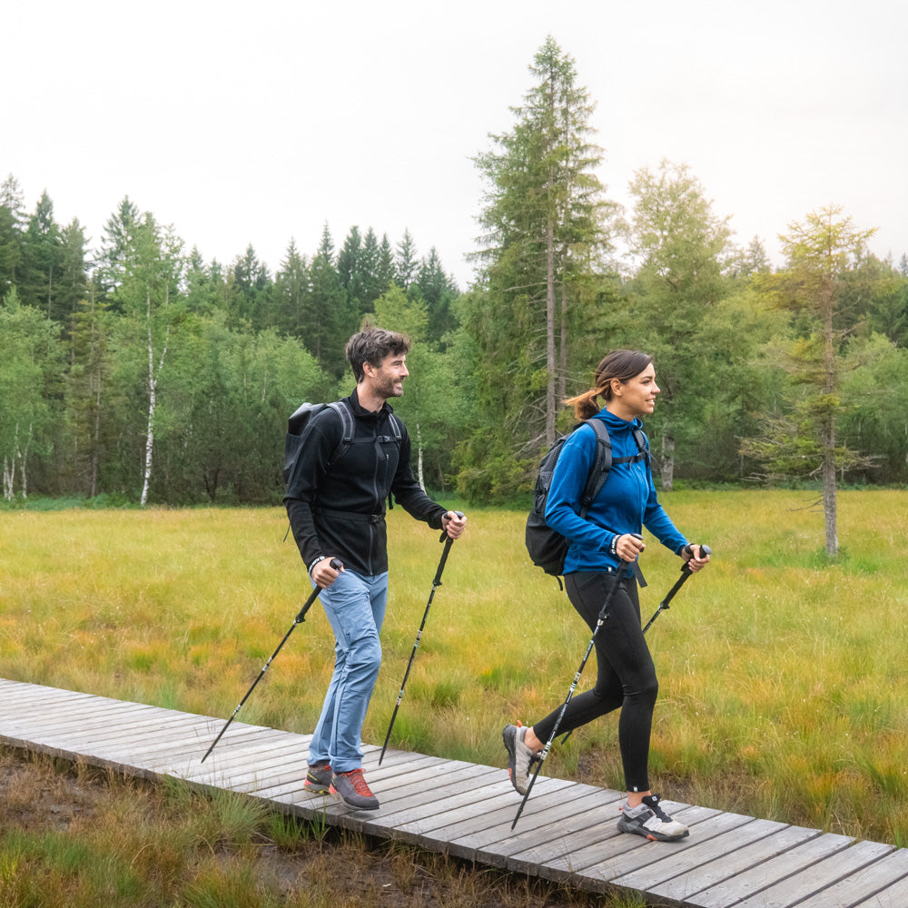 Zwei Personen wandern mit dem 100% Merino Sport Hoody mit Zip Damen - 230g/m2 durch ein Feuchtgebiet, nutzen Wanderstöcke und tragen Outdoor-Bekleidung und Rucksäcke.