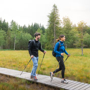 Zwei Personen wandern mit dem 100% Merino Sport Hoody mit Zip Damen - 230g/m2 durch ein Feuchtgebiet, nutzen Wanderstöcke und tragen Outdoor-Bekleidung und Rucksäcke.