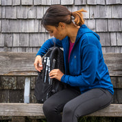 Frau mit blauem Merino Sport Hoody 230g/m², sitzt auf Bank, packt schwarze Trekkingstöcke ein. Ideal für Outdoor-Aktivitäten, atmungsaktiv und temperaturregulierend.