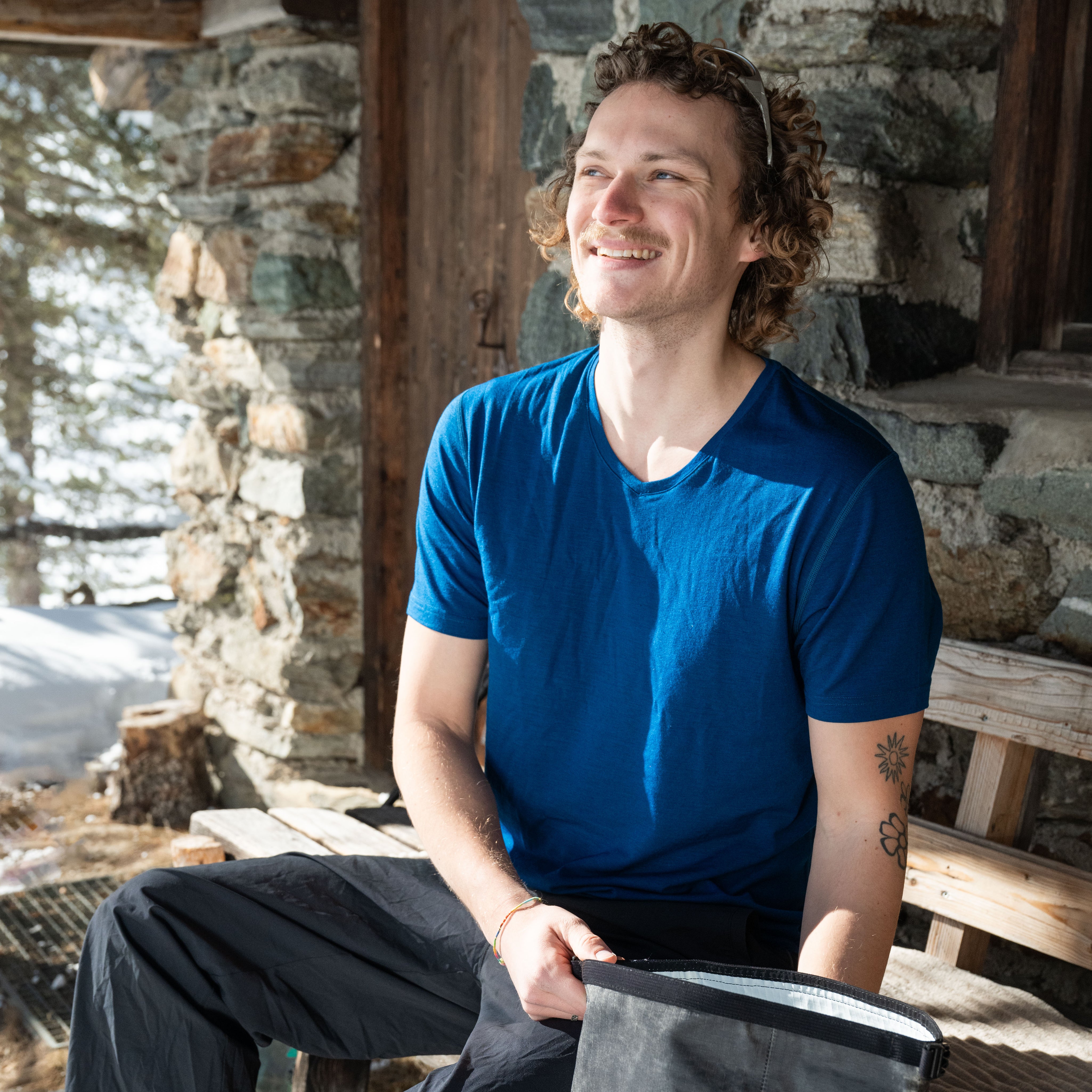 Herr sitzt auf einer Bank vor einer Hütte in blauem Merino V-Neck T-Shirt