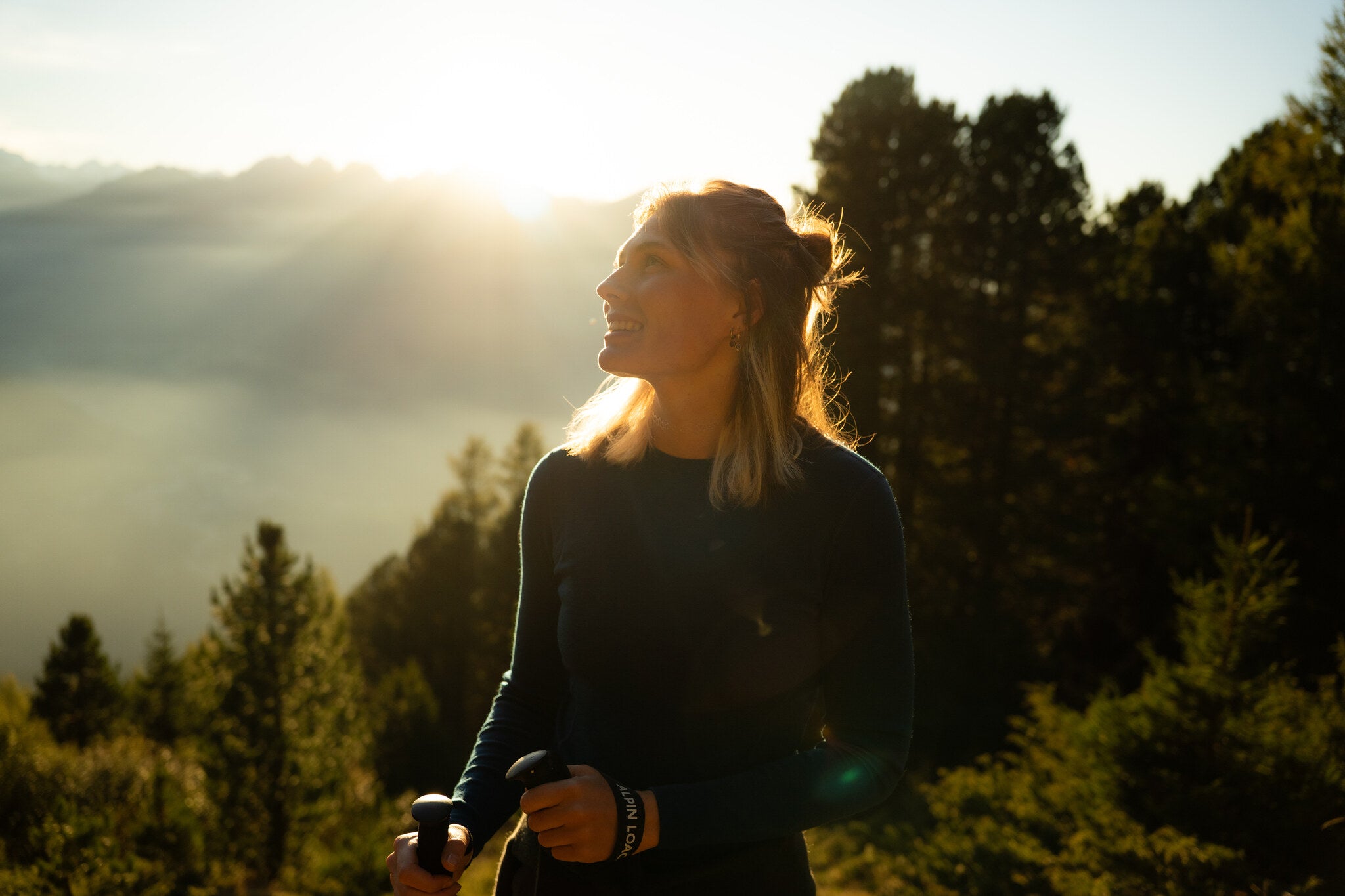Eine wandernde Frau steht am Berg in blauem Merino langarm Shirt mit Wanderstöcken in der Abendsonne am Berg