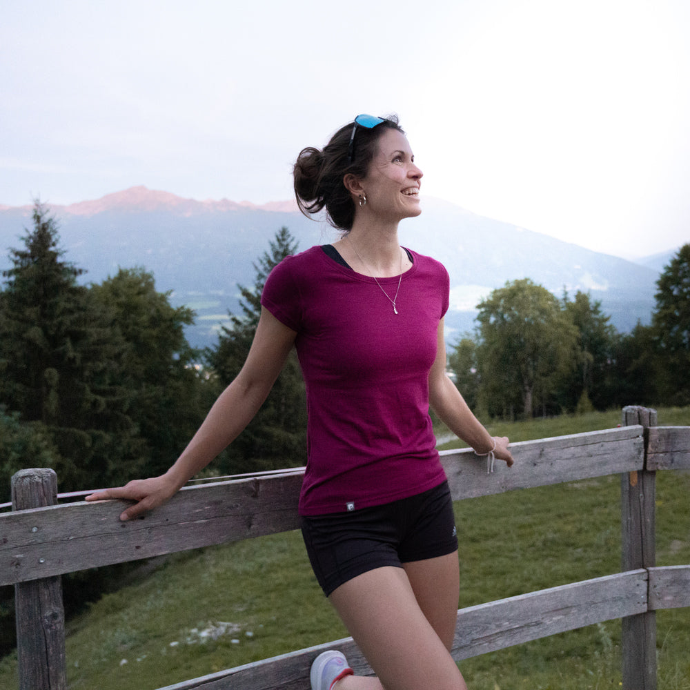Frau trägt ein sportliches Damen-T-Shirt in der Farbe Berry aus Merino-Coolmax-Materialmix, steht entspannt an einem Holzzaun mit Berglandschaft im Hintergrund – perfekt für Outdoor-Aktivitäten bei wechselnden Bedingungen.