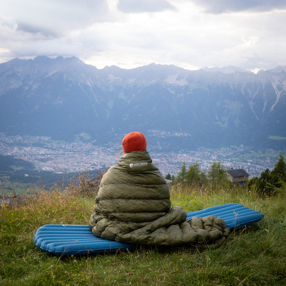 Frau sitzt im Schlafsack auf blauer Isomatte und schaut ins Tal