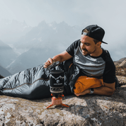 Camping Gaskocher Set: Mann entspannt auf Felsen, neben einem kompakten, ultraleichten Gaskocher, ideal für Outdoor-Abenteuer und effiziente Mahlzeitenzubereitung in der Natur.