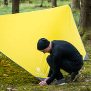 Mann kniet vor gelbem Zelt, schnürt Schuhe, neben Tarp 500 – das leichte Wingtarp für vielseitigen Outdoor-Einsatz und Schutz.