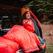 Frau im Auto mit dem 3 Jahreszeiten Schlafsack Bundle, hält einen roten Daunenschlafsack, ideal für Outdoor-Abenteuer und kühle Nächte.