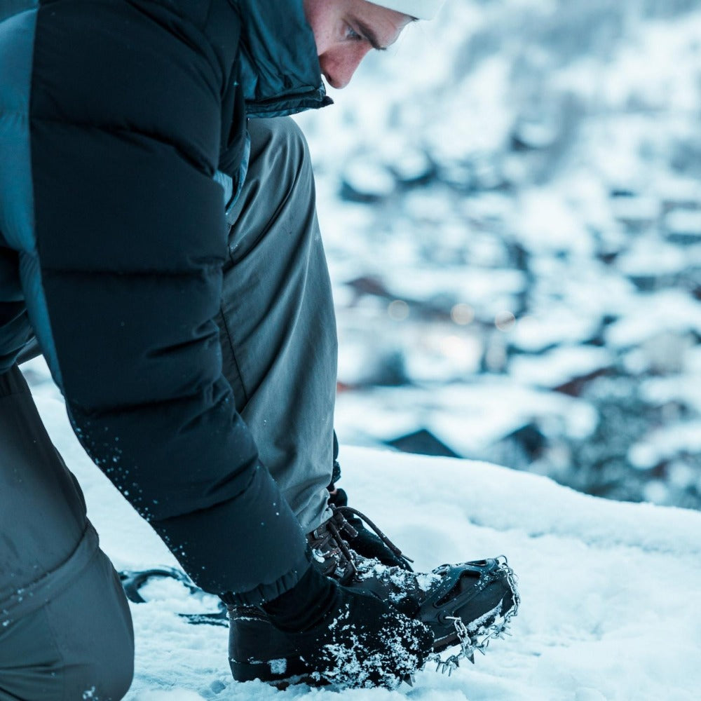 Mann bindet seine Schuhe im Schnee, trägt Chain Pro 18 Grödel mit 18 Spikes für sicheren Halt auf winterlichen Pfaden. Perfekt für Trekking und Bergtouren.