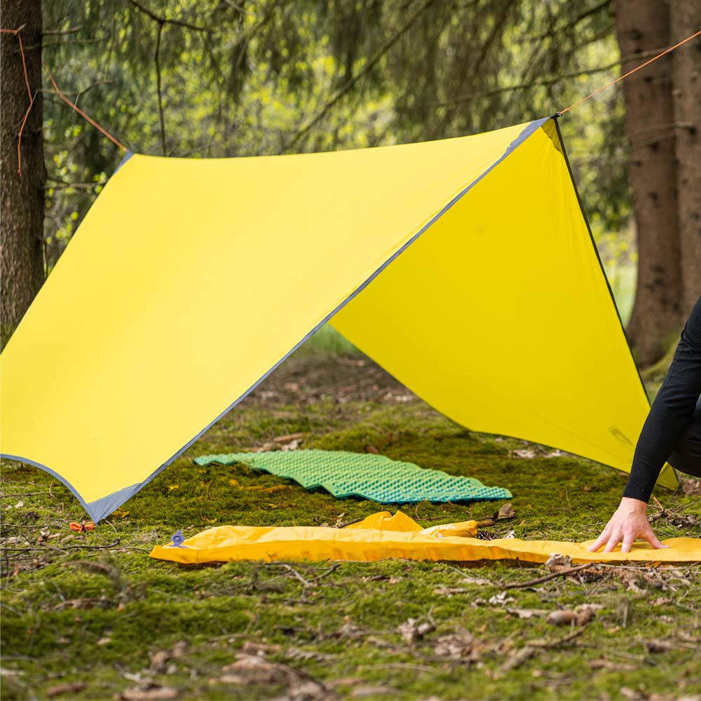 Person neben gelbem Tarp 500 - das leichte Wingtarp, auf Gras aufgebaut, ideal für Outdoor-Abenteuer als vielseitiger Schutz bei Wetterbedingungen.