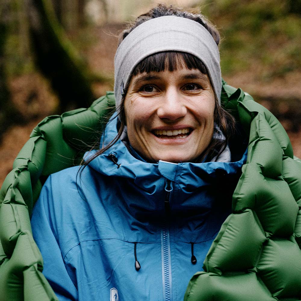 Frau in einer blauen Outdoor-Jacke, die die ultraleichte Isomatte - 465g von alpinloacker.com für Camping präsentiert, mit Fokus auf Schlafkomfort und Nachhaltigkeit.