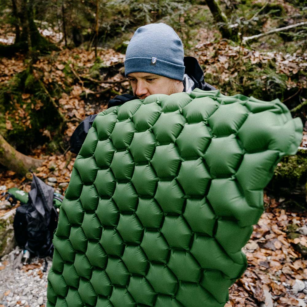 Mann mit blauer Mütze hält ultraleichte Isomatte - 465g, ideal für umweltfreundliches Camping, aus recyceltem 20D RipStop Nylon, im Wald.